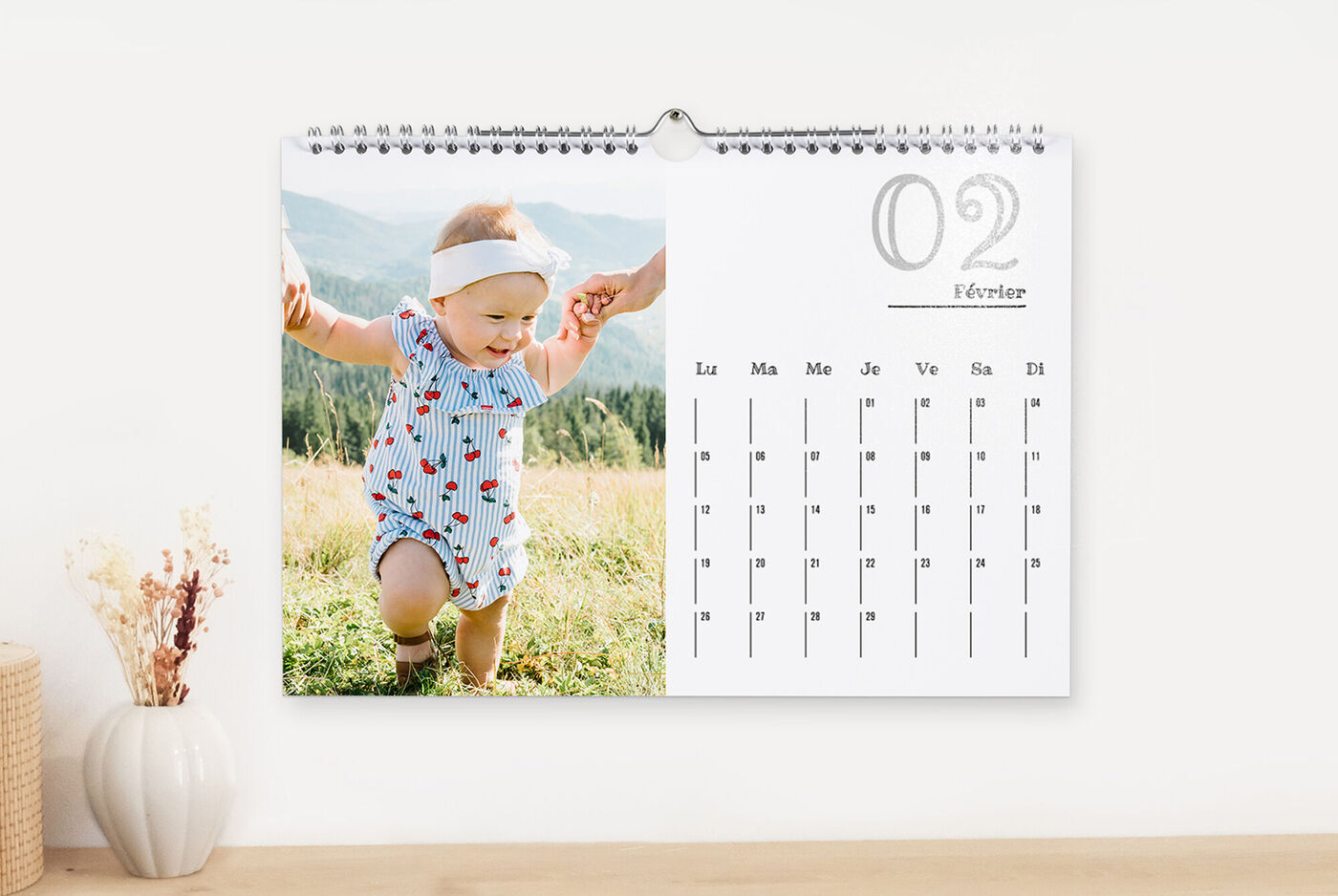 Un petit enfant en blouse colorée marche sur une pelouse, tenu par des adultes. Des collines vertes en arrière-plan.