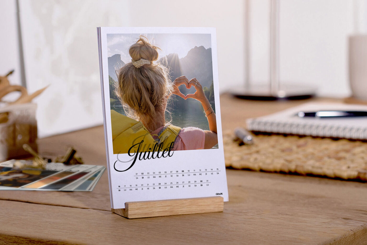 Une fille fait un geste de cœur devant une table. En arrière-plan, un calendrier. CEWE calendrier photo.