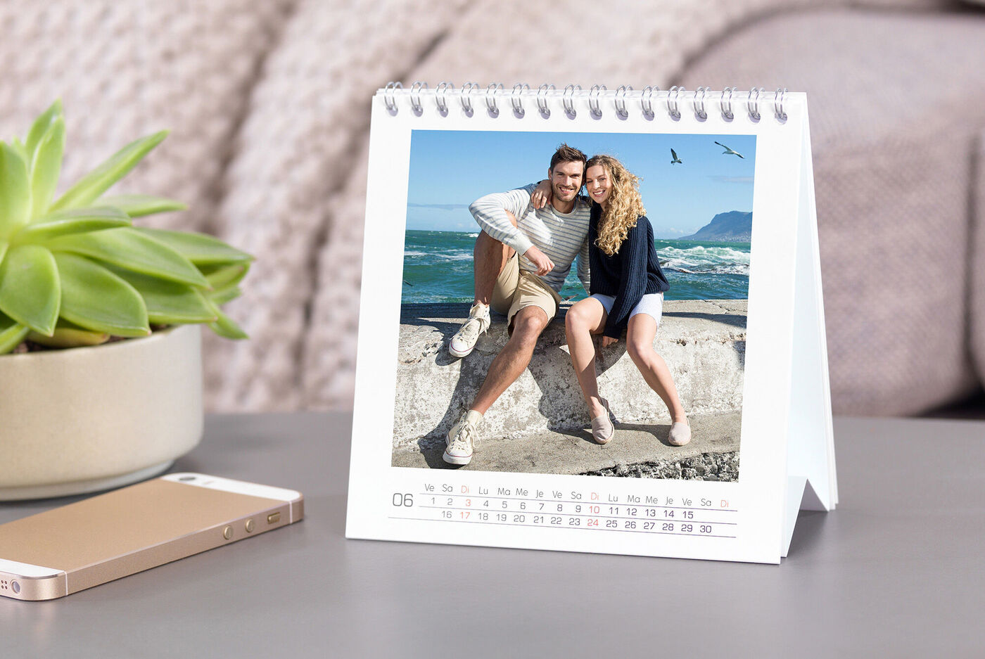 Un couple amoureux est assis sur un mur à la plage, avec des vagues en arrière-plan. Un calendrier photo est sur la table.