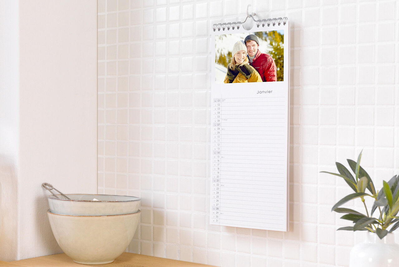 Un calendrier photo CEWE pend au mur. En haut, une photo joyeuse d'un couple dans la neige. En bas, les feuilles du calendrier avec un champ libre pour janvier.