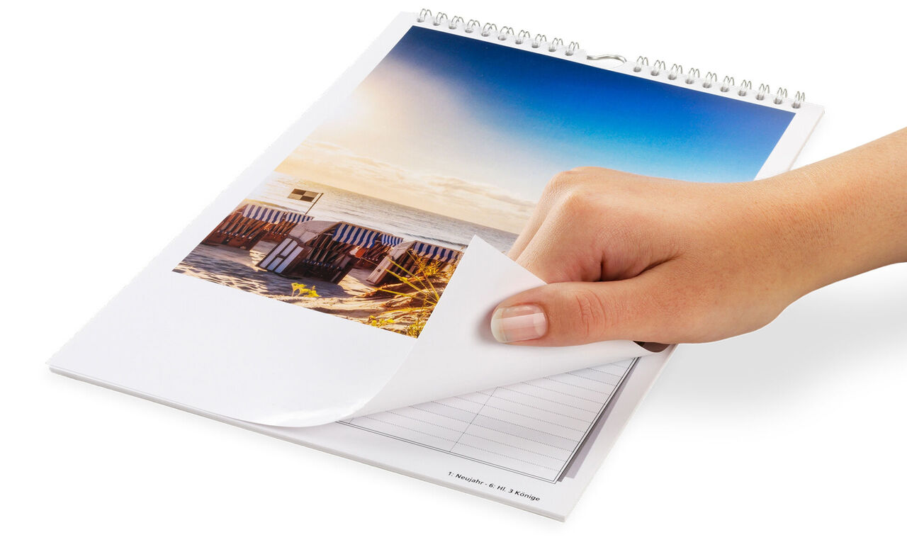 Une main feuillette un CEWE calendrier photo avec un beau motif de plage. Le ciel est visible en arrière-plan.