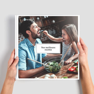Deux mains tiennent un livre photo de recettes. En arrière-plan, une cuisine confortable.