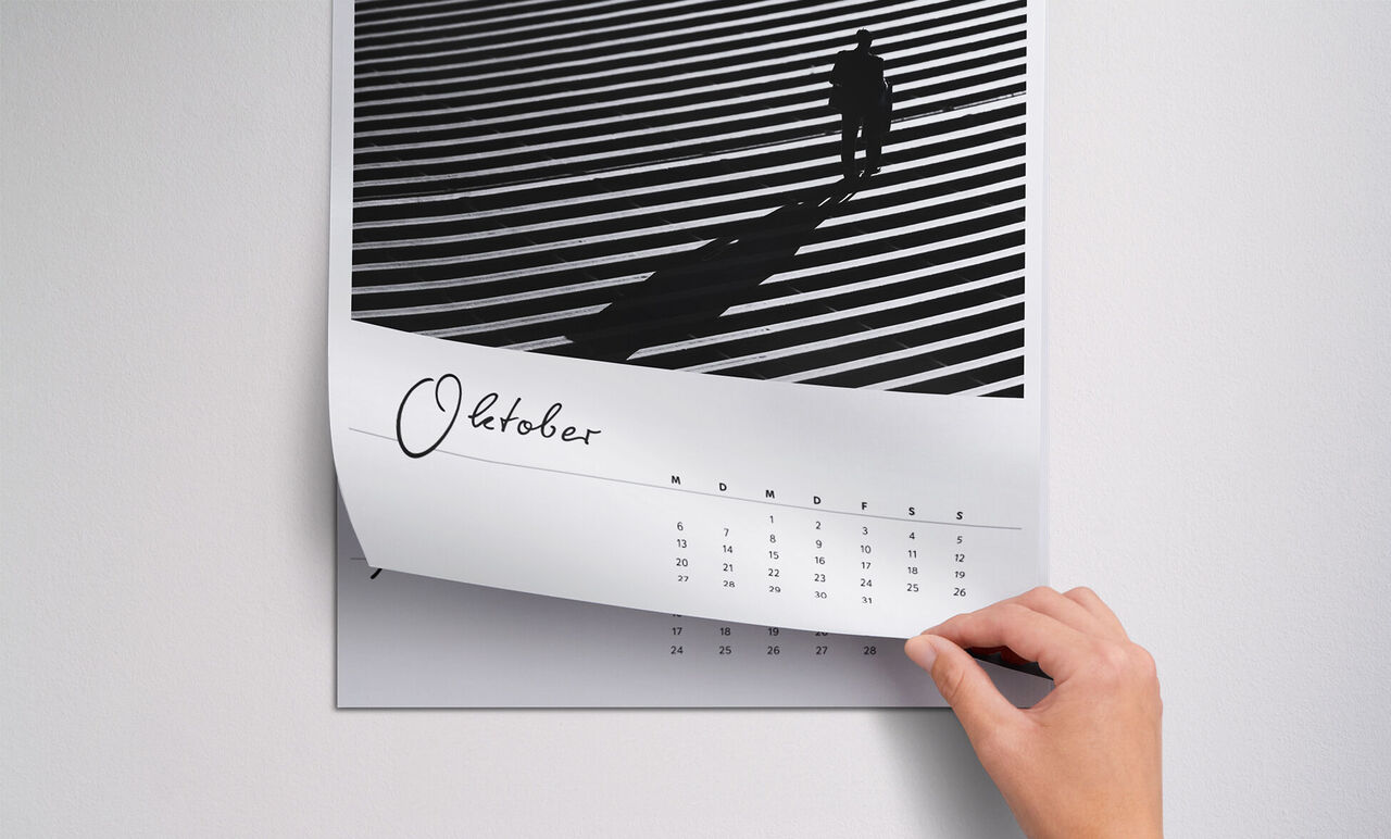 Une main feuillette un calendrier photo avec des lignes noir et blanc et une personne dans l'ombre. Le mois d'octobre est visible.