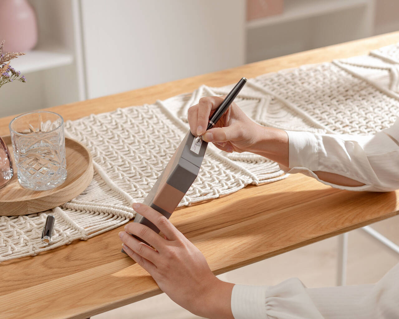 Une personne tient un carnet gris et écrit avec un stylo noir. La table est en bois et comporte des éléments décoratifs.