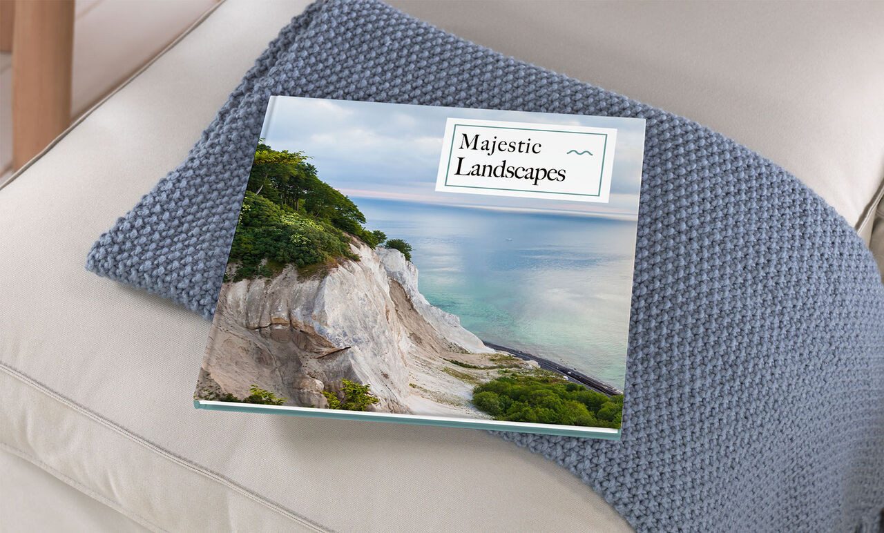 Un album CEWE ouvert intitulé "Majestic Landscapes" repose sur un canapé beige, entouré d'un coussin bleu.