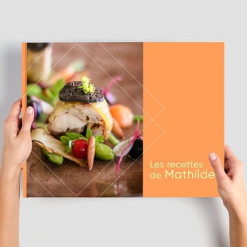 Une personne tient un livre photo CEWE intitulé "Les recettes de Mathilde" avec un plat de poisson.